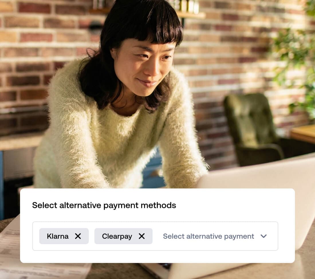 A woman leaning over a laptop in a kitchen setting, looking at the screen. Overlaid on the image is a user interface window titled "Select alternative payment methods" showing selected tags for "Klarna" and "Afterpay" next to a dropdown menu.