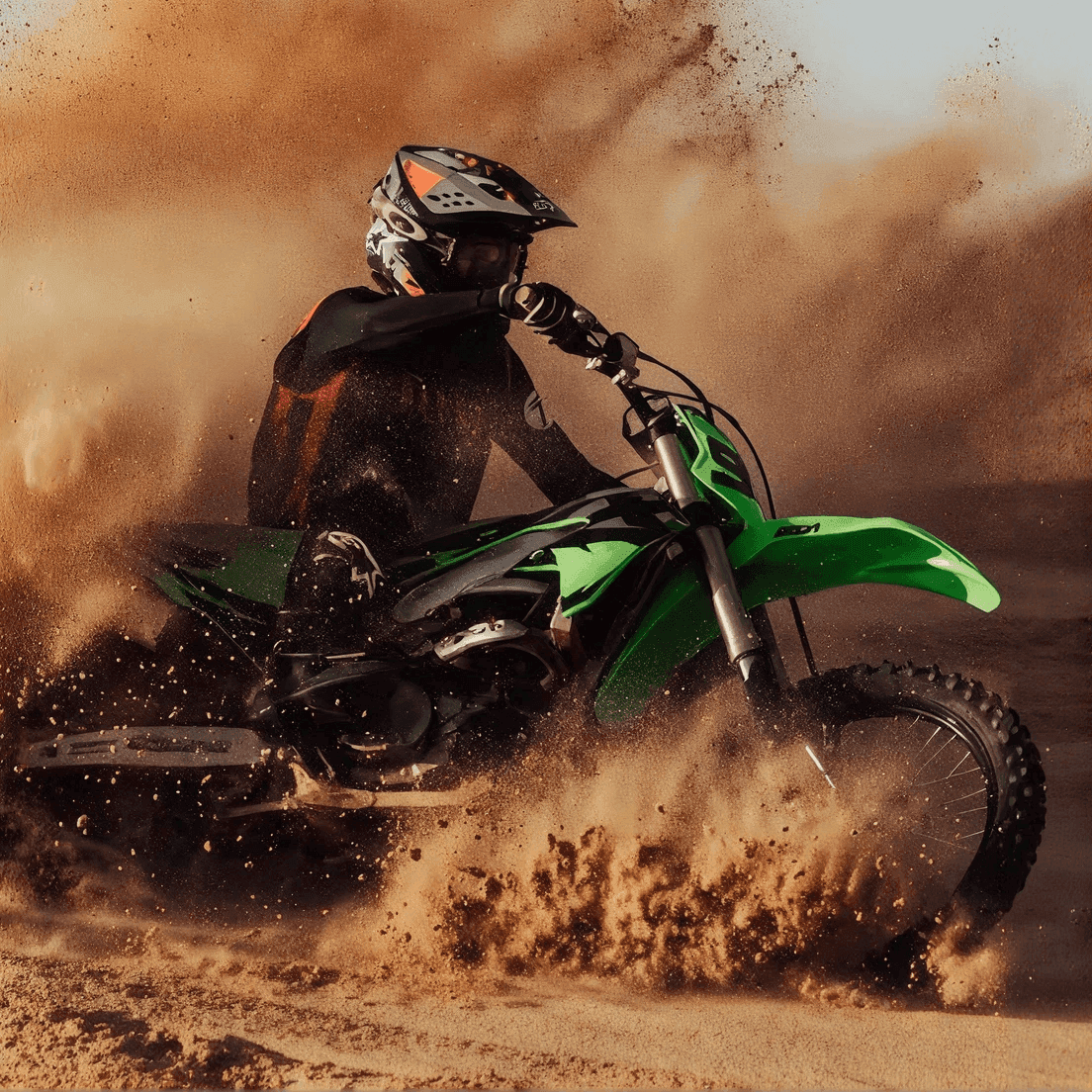 Motocross rider kicking up sand and dust on a green off-road dirt bike in a sharp turn.